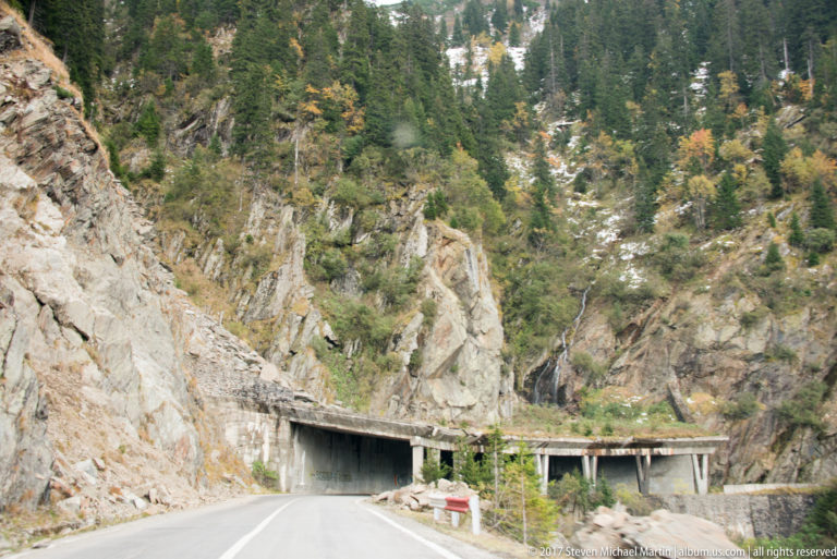 Road entering concrete protective galleries beneath fractured rock and conifer forest in the mountains.