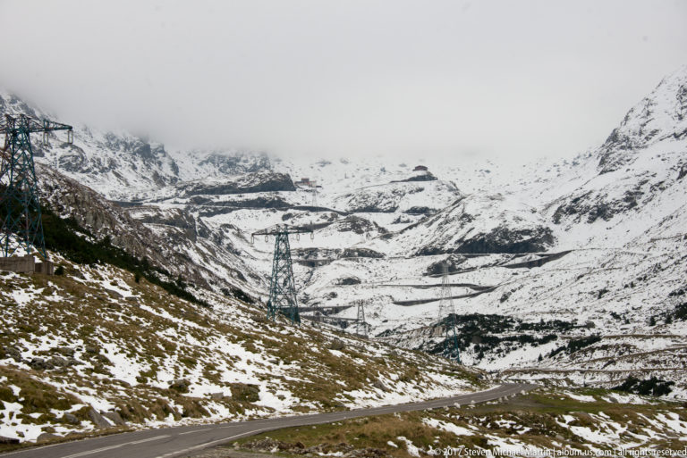 Snow-covered mountain basin with looping road switchbacks and several electrical pylons.
