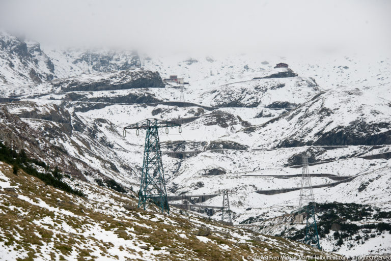 Electrical pylons stand before dark road switchbacks crossing a snow-covered mountain slope.