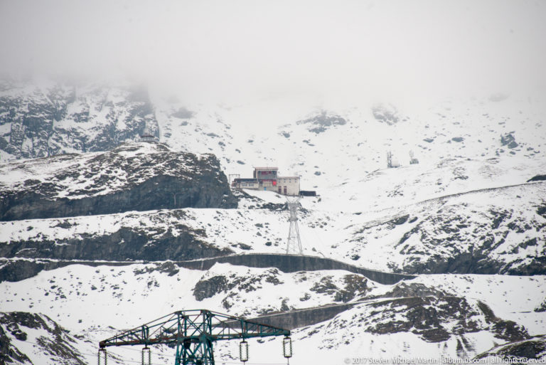 Rectangular building on a snowy mountain bench above a steel electrical pylon beneath low cloud.
