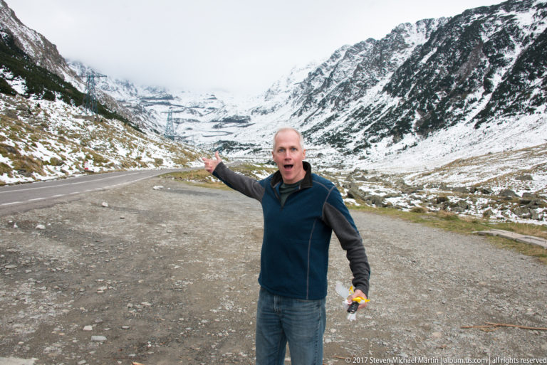 Visitor gesturing toward the Transfăgărășan in a broad snow-covered mountain basin.