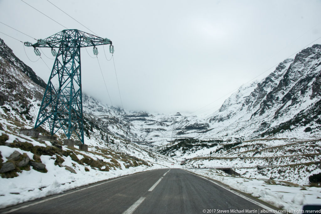 Dark two-lane road through snow-covered mountain slopes beneath a steel electrical pylon and low cloud.