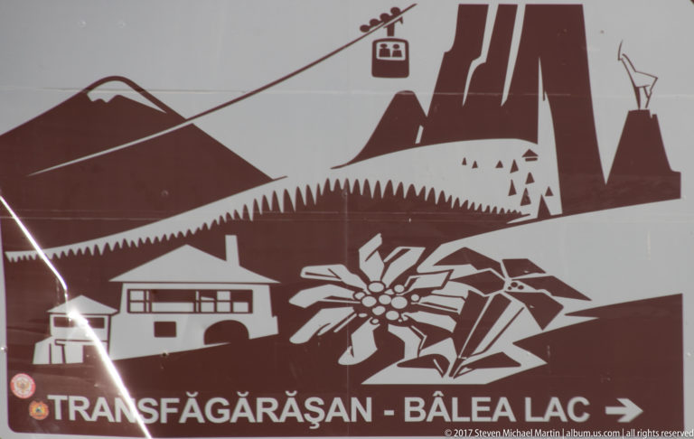 Close-up of the Transfăgărășan–Bâlea Lac sign with mountain, cable-car, chalet, forest, and edelweiss graphics.