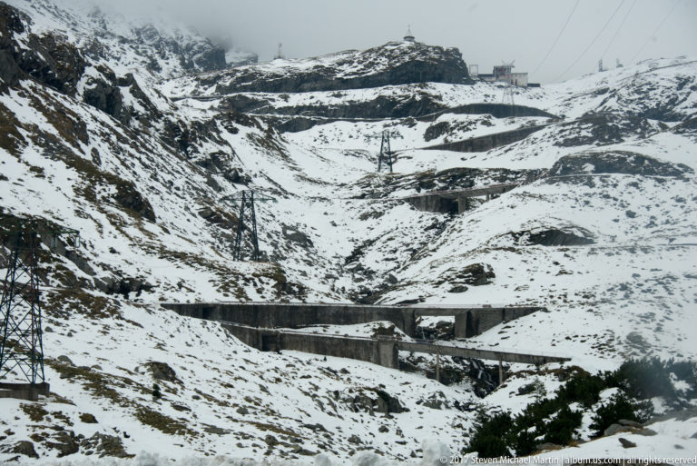 Snow-covered slope crossed by retaining walls, road terraces, electrical pylons, and small ridge-top structures.