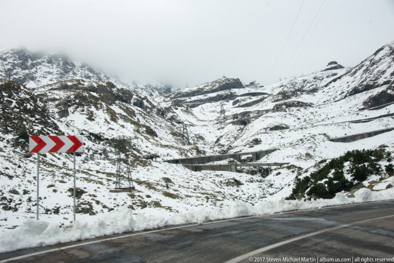 Sharp snow-lined mountain-road bend with red-and-white chevron signs and terraced slopes beyond.