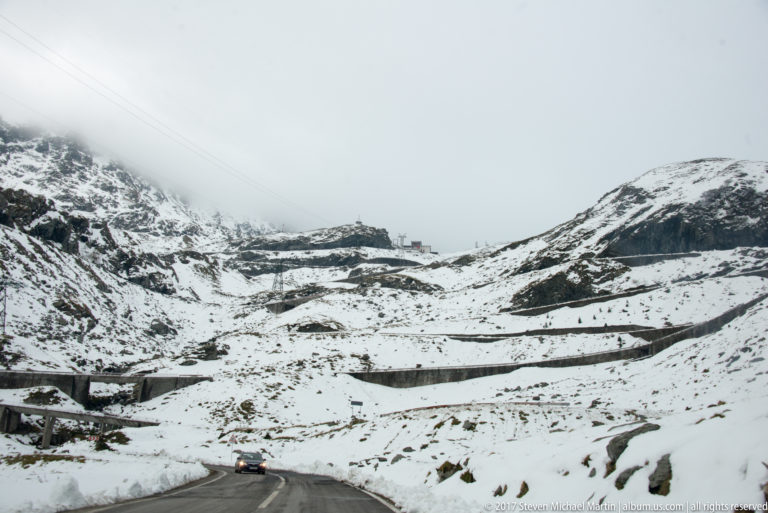 Car driving on the Transfăgărășan beneath snowy switchback slopes and low cloud.