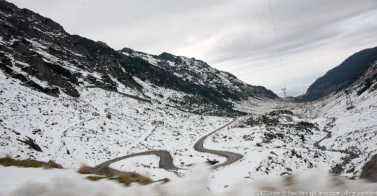 Elevated view of dark Transfăgărășan hairpin curves winding through a snow-covered mountain valley.