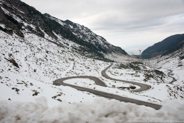 Multiple Transfăgărășan hairpin curves descending through a snowy alpine trough between dark mountain slopes.