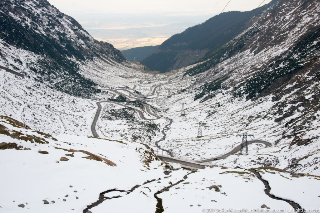 High view down a snowy valley with winding Transfăgărășan road, stream channels, and electrical pylons.