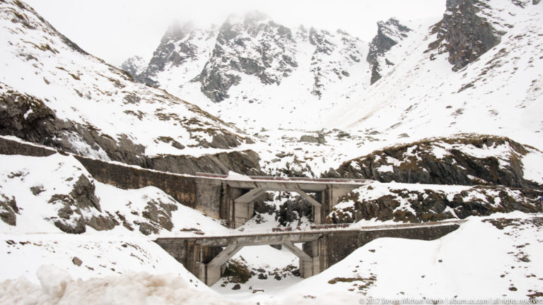 Concrete bridge and retaining structures crossing a snow-filled rocky gully in the high mountains.