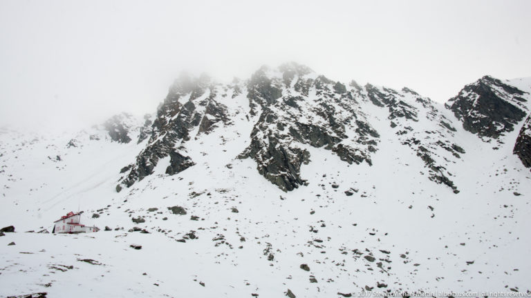 Jagged dark rock rising through deep snow beneath low cloud, with small buildings at the base of the slope.