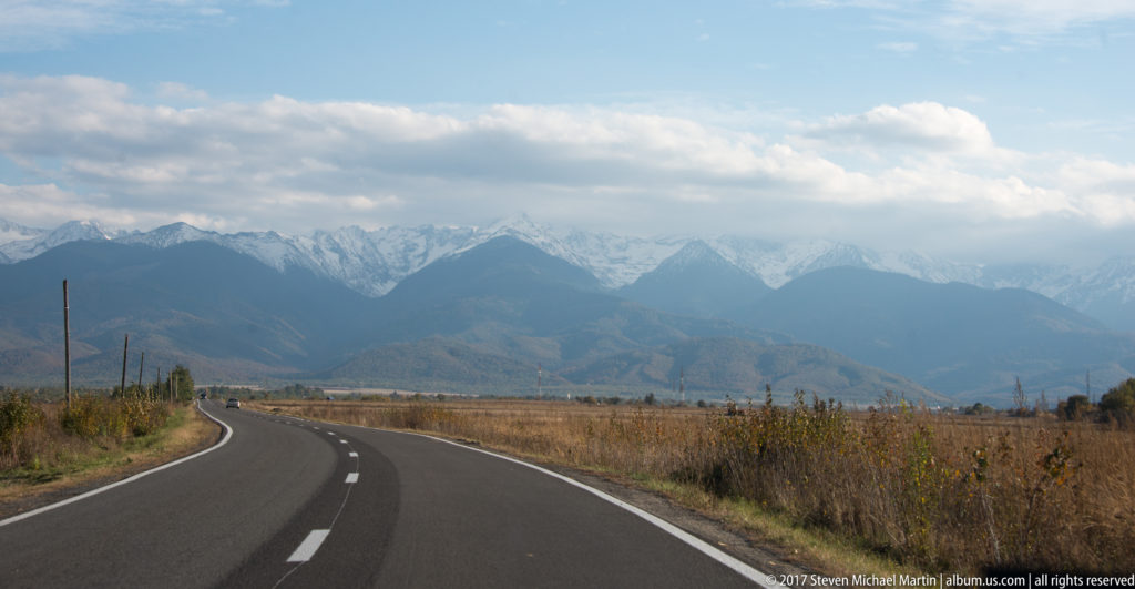Two-lane mountain road curving toward jagged Făgăraș peaks beneath broken clouds and blue sky.