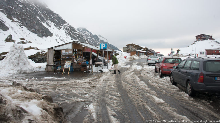 Cars, visitors, and wooden roadside stalls in a slushy snow-covered parking area beneath mountain slopes.