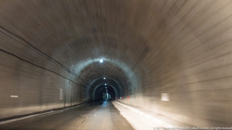 Long concrete road tunnel with repeating ceiling lights and a bright opening in the distance.