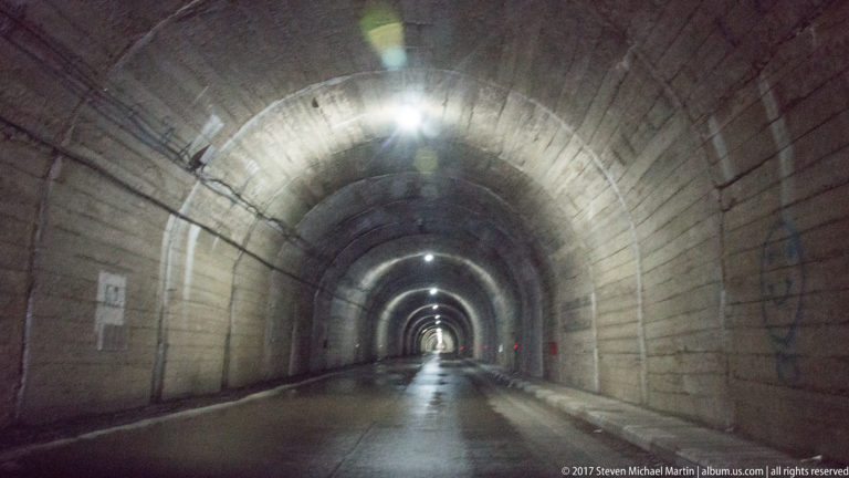 Centered view through an arched concrete tunnel with wet pavement, lights, and a bright distant exit.