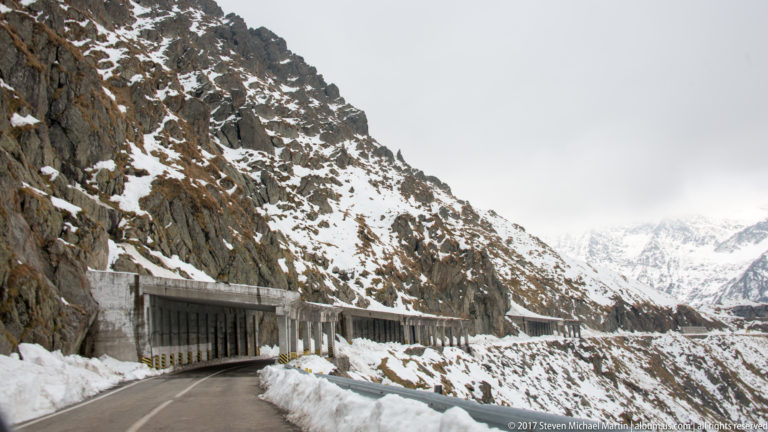 Concrete road gallery at the base of a snowy rock cliff with mountains beyond.