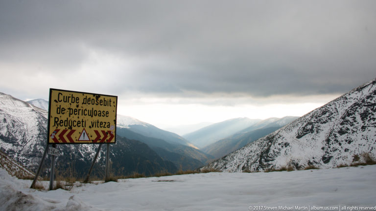 Romanian warning sign for dangerous curves and reduced speed beside a snowy mountain valley.