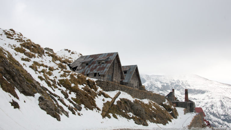 Weathered steep-roofed mountain buildings on a snowy hillside above the road.