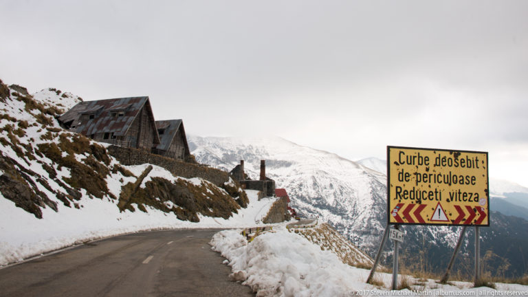 Dangerous-curves warning sign beside a snowy mountain road and weathered buildings.