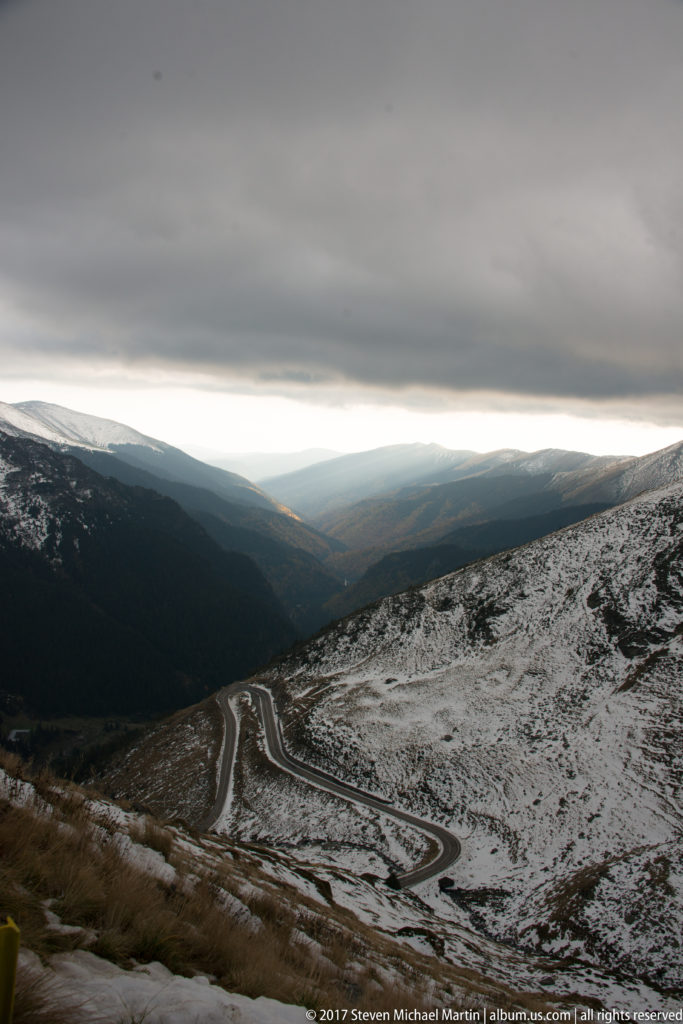 Dark road switchbacks descending across patchy snow toward a distant mountain valley.