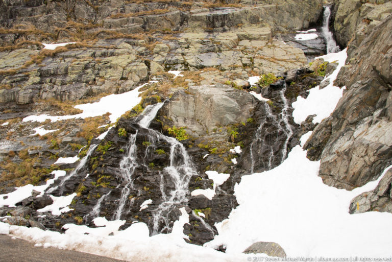 Small mountain cascade flowing over dark bedrock between patches of melting snow.