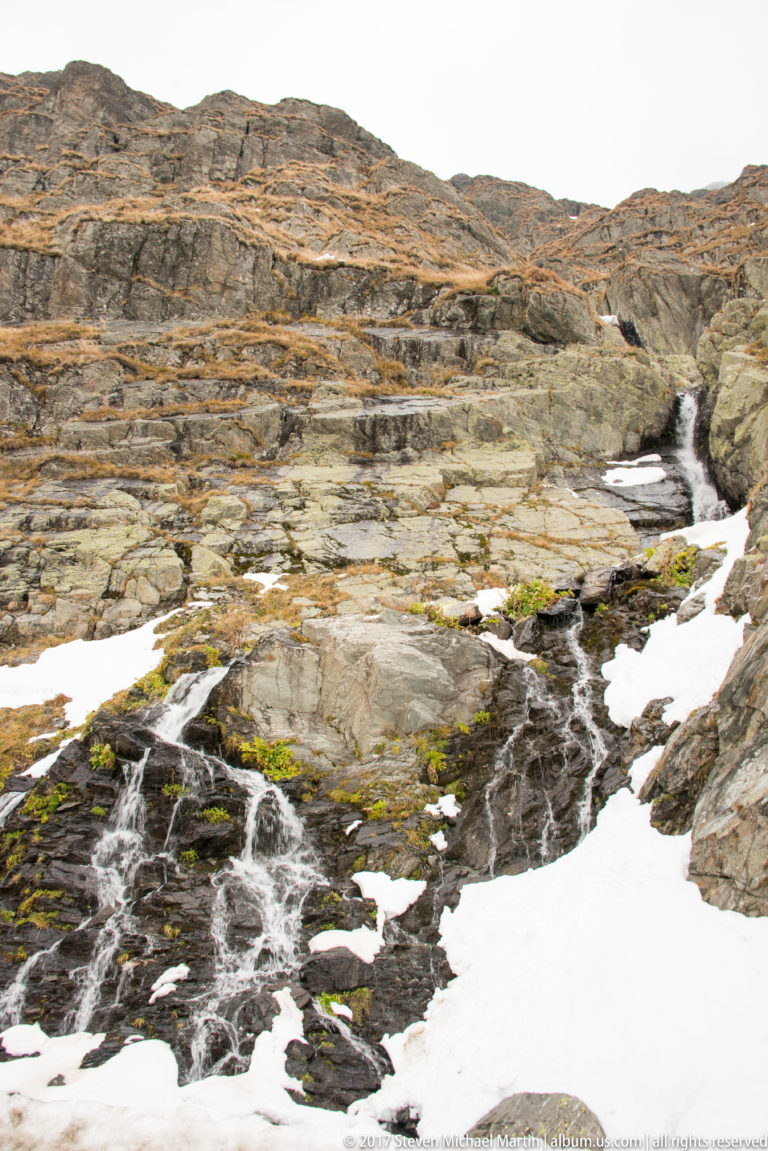 Snowmelt stream spreading across layered rock and dropping through a narrow alpine channel.