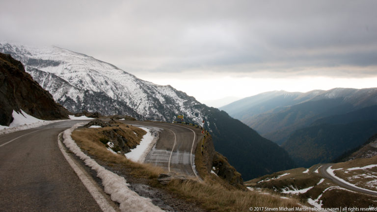 Curving mountain road above a deep valley with patchy snow and distant ridges.