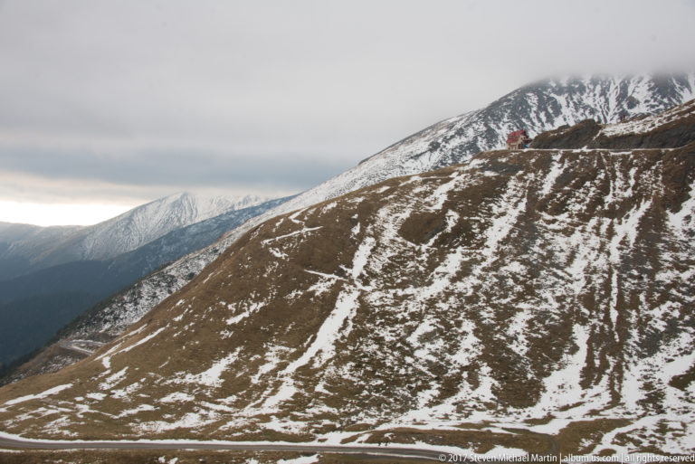 Broad brown mountain slope streaked with snow and a narrow road line beneath cloud.
