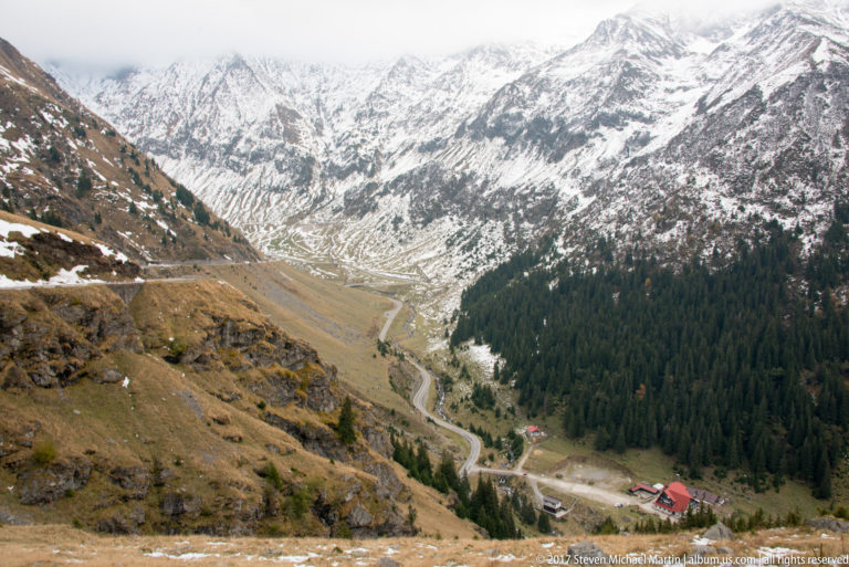 High view into a mountain valley with winding road, conifer forest, and snow-covered upper slopes.
