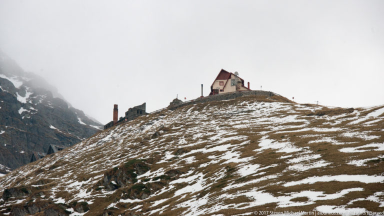 Small steep-roofed house on a snow-patched mountain ridge beneath misty cliffs.