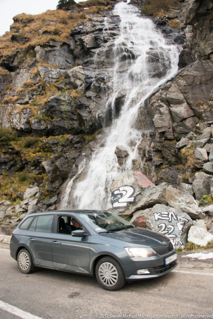 Gray station wagon passing a roadside cascade flowing over graffiti-marked rocks.