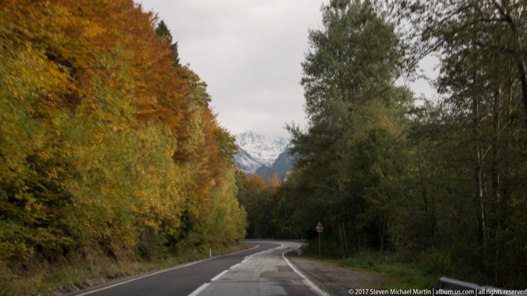 Wet mountain road between autumn trees, with a snow-dusted peak visible ahead beneath gray sky.