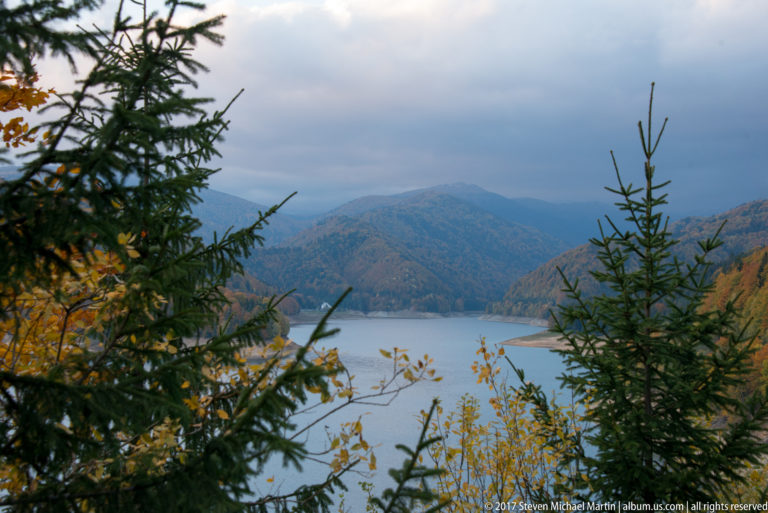 Mountain reservoir and forested ridges seen through spruce trees and autumn foliage beneath an overcast sky.