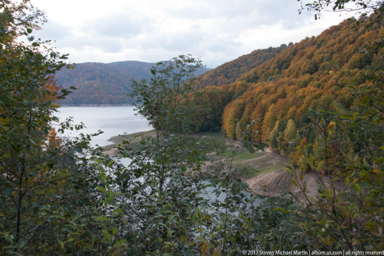 Reservoir shoreline curves below a steep hillside covered with orange, gold, and green autumn forest.