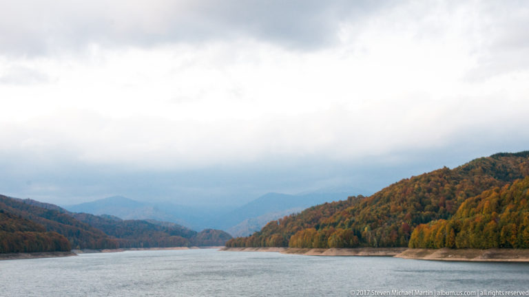 Long reservoir extends between autumn-covered hills toward hazy blue mountain ridges under low clouds.