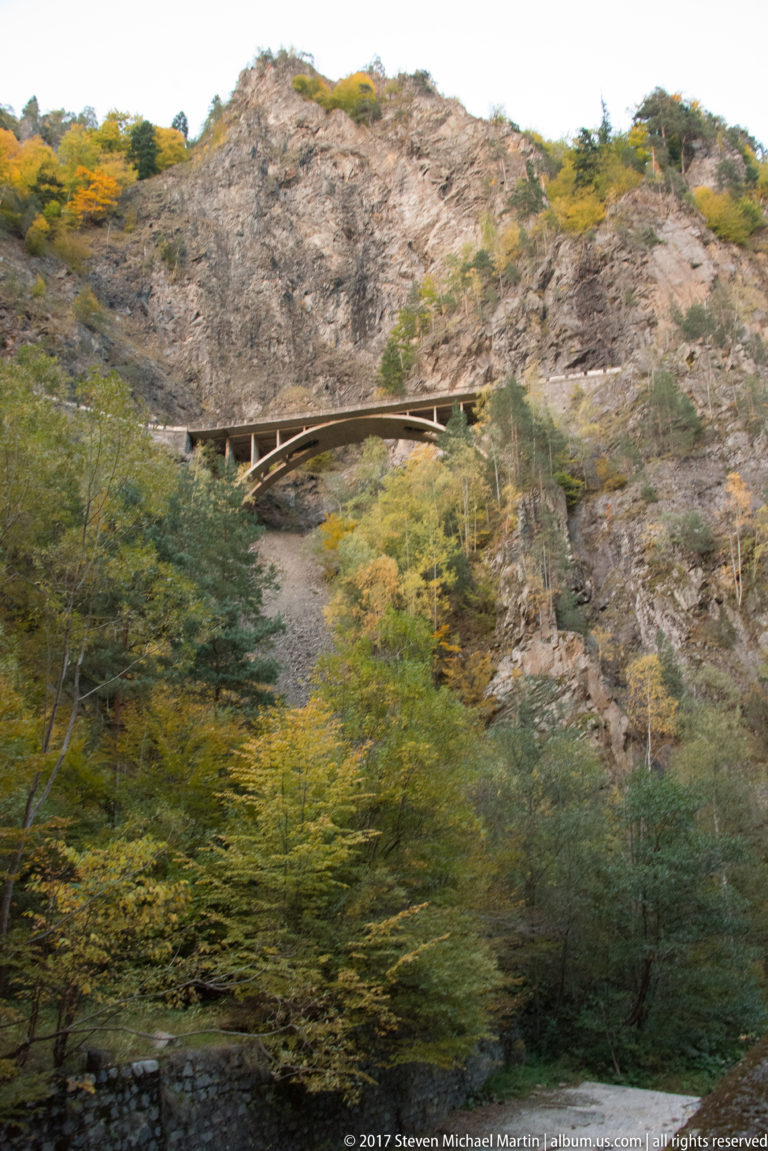 Narrow wooden structure crosses a fractured rocky slope above dense green and autumn-colored woodland.