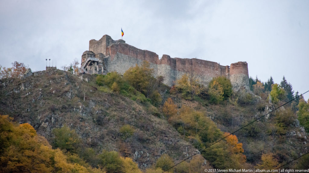 Ruined hilltop fortress with long stone walls and Romanian flag rises above a steep autumn-covered slope.