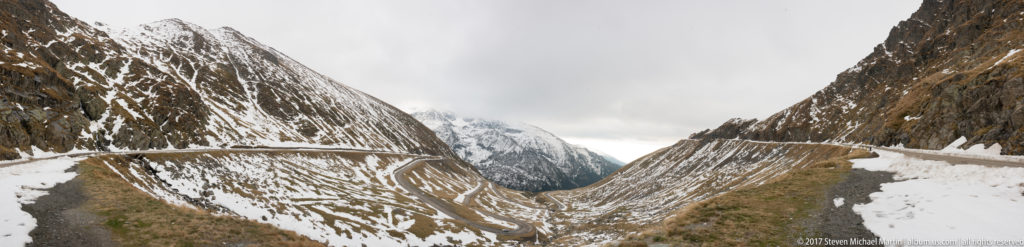 Snow-lined Transfagarasan Road crosses steep mountain slopes toward cloud-covered peaks in a narrow alpine valley.