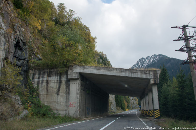 Concrete protective road gallery beneath a rocky slope, with striped hazard markings and a mountain beyond.