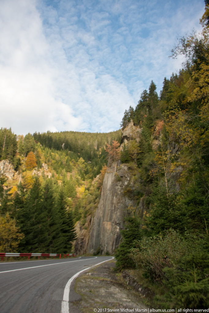 Curving mountain road beside exposed rock face and mixed autumn forest beneath a blue-and-cloudy sky.
