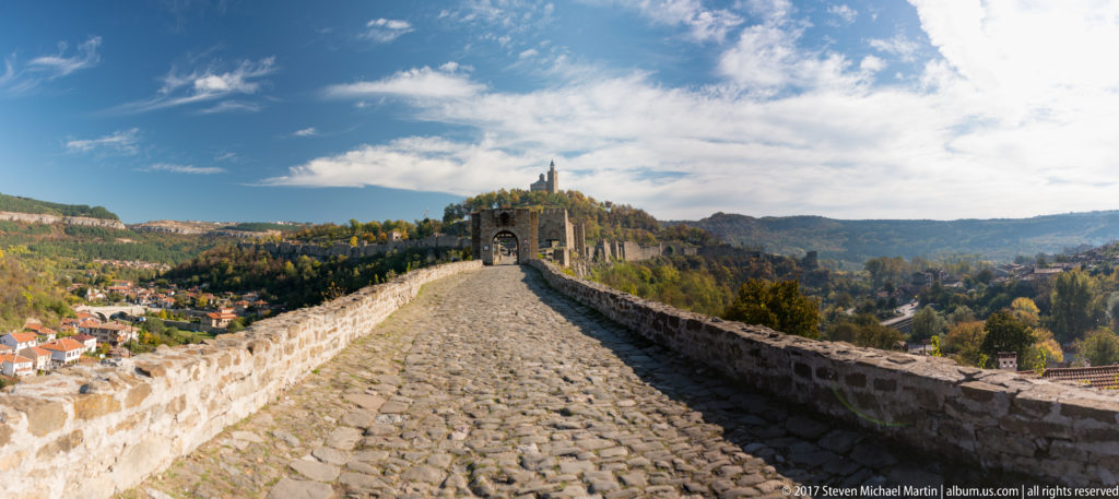 Cobblestone rampart walk between stone parapets leading toward an arched fortified gateway above Veliko Tarnovo.