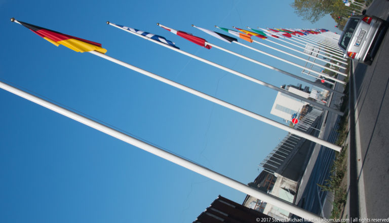 Diagonal row of tall flagpoles with international flags against a clear blue sky.