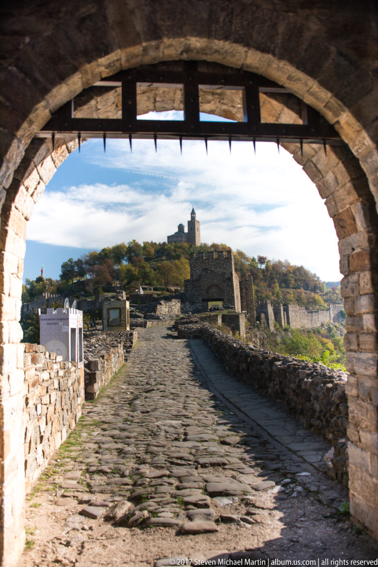 View through a stone fortress arch toward a cobbled road, ruins, and distant hilltop tower.