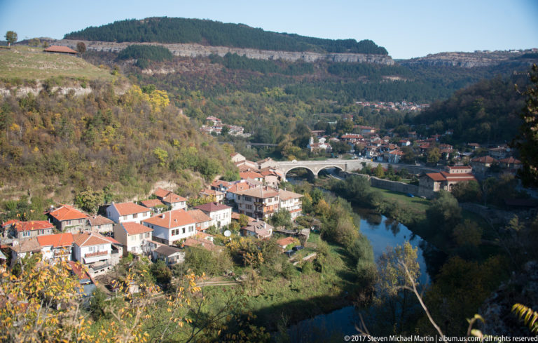 Red-roofed houses beside a winding river and multi-arched bridge in a steep wooded valley.