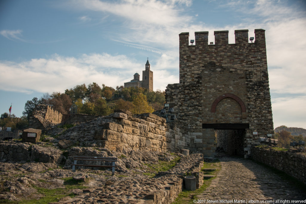 Crenellated rectangular stone gate tower amid fortress ruins, with a hilltop tower in the background.