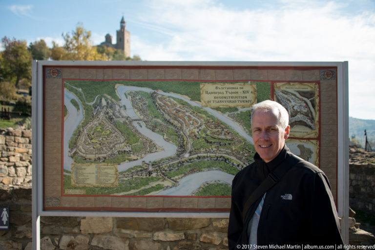 Smiling visitor standing beside a large illustrated fortress reconstruction map with a tower behind.