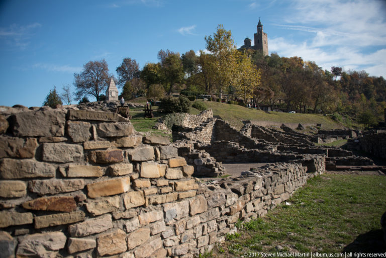 Low stone foundations and ruined walls stepping up a grassy fortress hillside toward a distant tower.