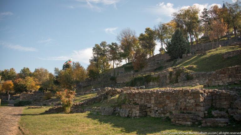 Stone foundations and terraced ruins across a grassy autumn hillside, with a Bulgarian flag in the distance.