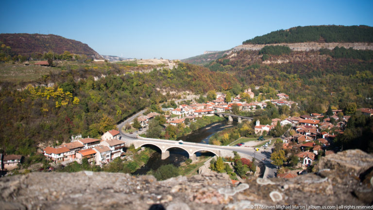 High view over a multi-arched bridge, river, and red-roofed neighborhood between steep wooded hills.
