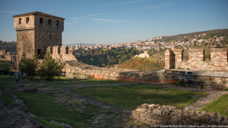 Square stone defensive tower beside crenellated fortress walls, with Veliko Tarnovo on the distant hillside.